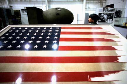 IMAGE: A tattered US flag in the foreground, a man looking at a bomb in the background